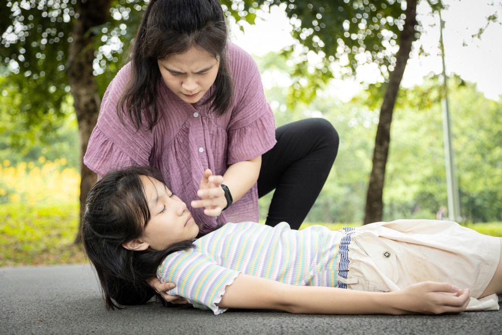 A woman checks the pulse of a young girl lying on the ground in a park, appearing concerned about a potential youth heart attack